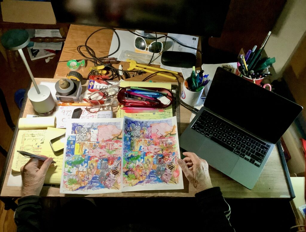 A photo of Luis's desk with his hands braced on it, the left holding a pen. Two color ink drawings lie on the table, along with a yellow legal pad, glasses, more colored pens, and a dark laptop computer.
